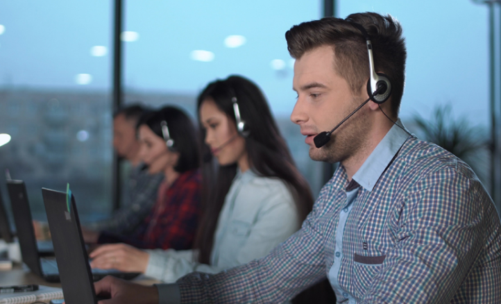 A row of four people sits at a table all wearing headsets and looking at laptops.